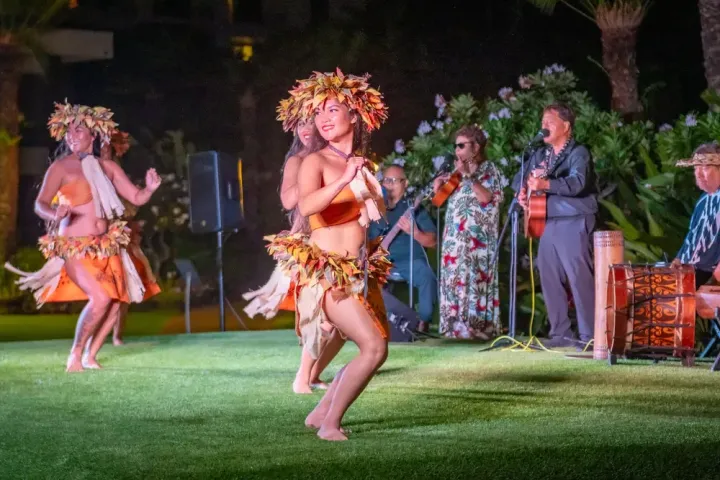 Hula dancers perform in grass, musicians play in the background.