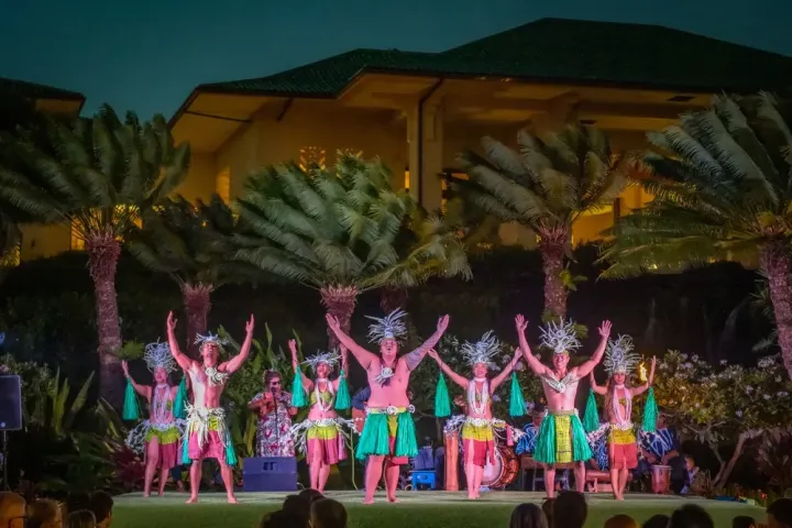 Dancers in vibrant costumes perform outdoors, with palm trees and a building in the background.