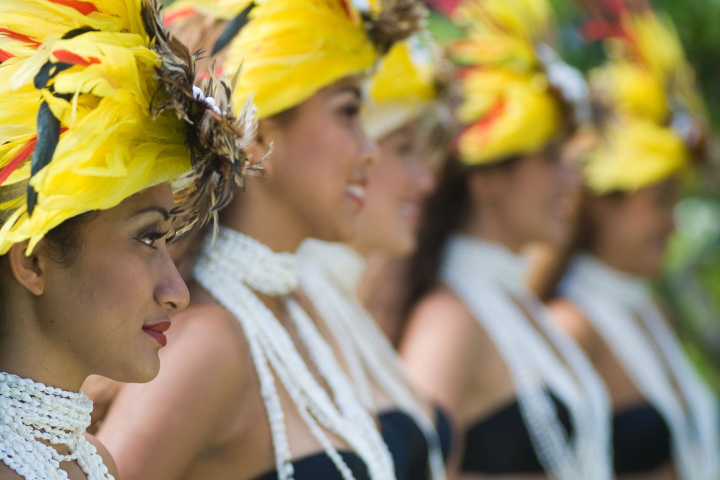 Women in colorful feather headdresses and pearl necklaces, standing in line, smiling.