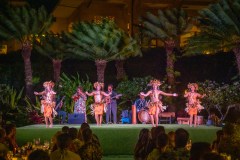 Dancers in traditional attire perform on a stage with musicians and palm trees in the background.