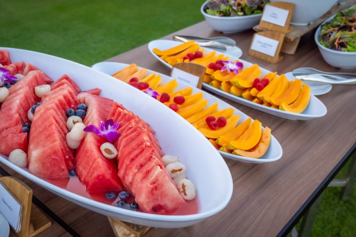 Plates of sliced watermelon and papaya with berries and flowers on a wooden table outdoors.