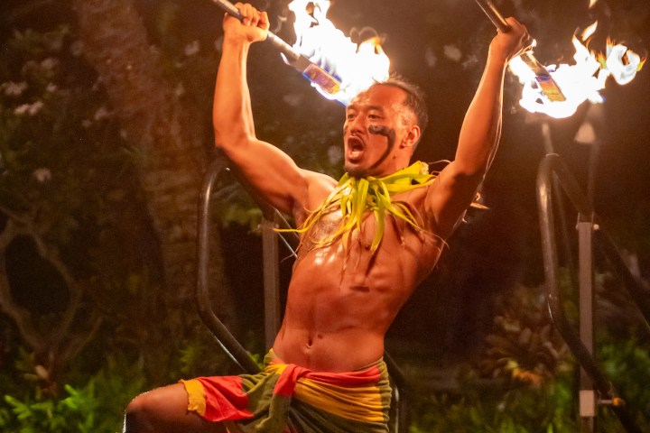 Performer kneeling with flaming torches in both hands during a fire dance show.