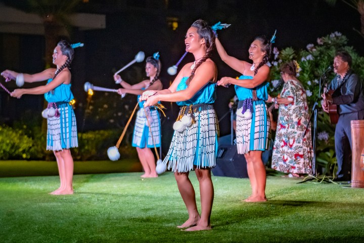 Dancers in traditional attire perform with poi balls on a grass stage at night.