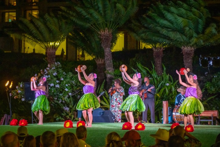 Hula dancers perform in colorful outfits on an outdoor stage, surrounded by lush greenery.
