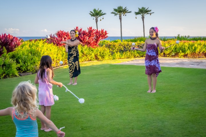 Two women teach poi dancing to children on a grassy area with tropical plants and ocean view.